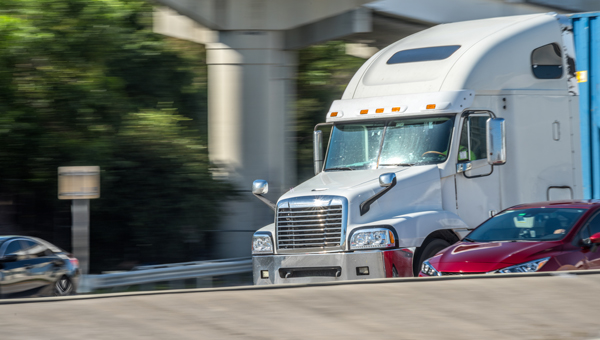 White tractor trailer rig driving under highway underpass with red car on its right